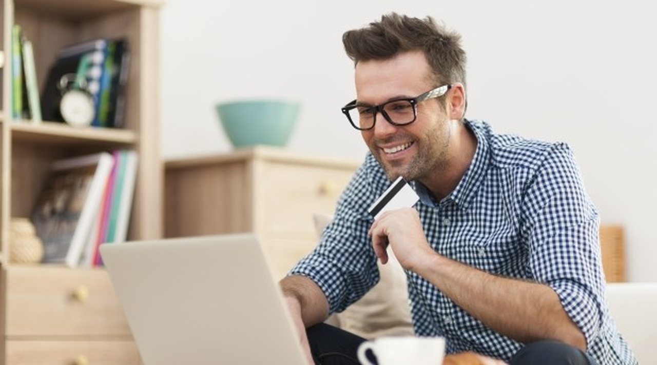 o-que-e-preciso-para-se-tornar-freelancer - homem sorrindo em frente ao computador com cartão na mão