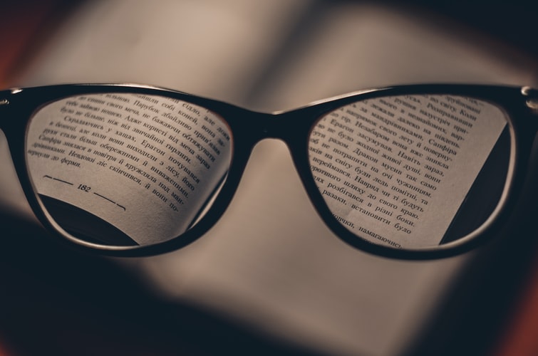 A imagem mostra um óculos. A lente desses óculos está focando em palavras de um livro.